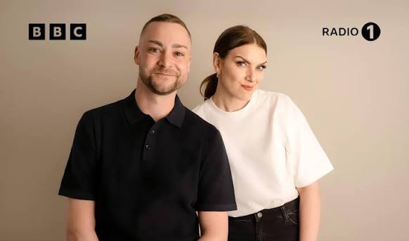 two people, a man and woman in front of a plain background, looking at the camera