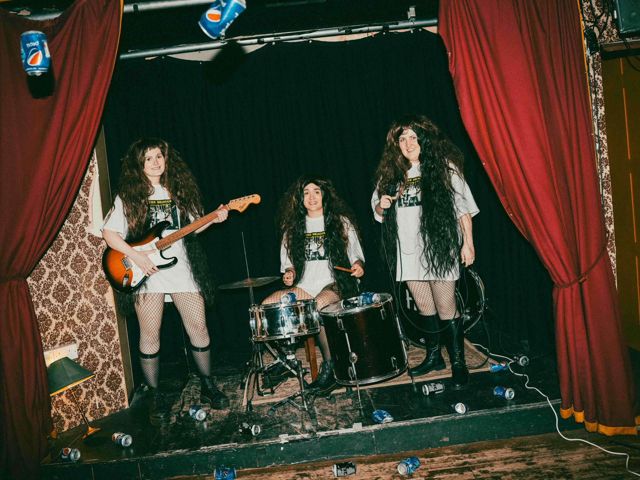 three people stood on a small stage with long black wigs, red curtains around them, with one on a drum kit, one holding a guitar and another holding a microphone