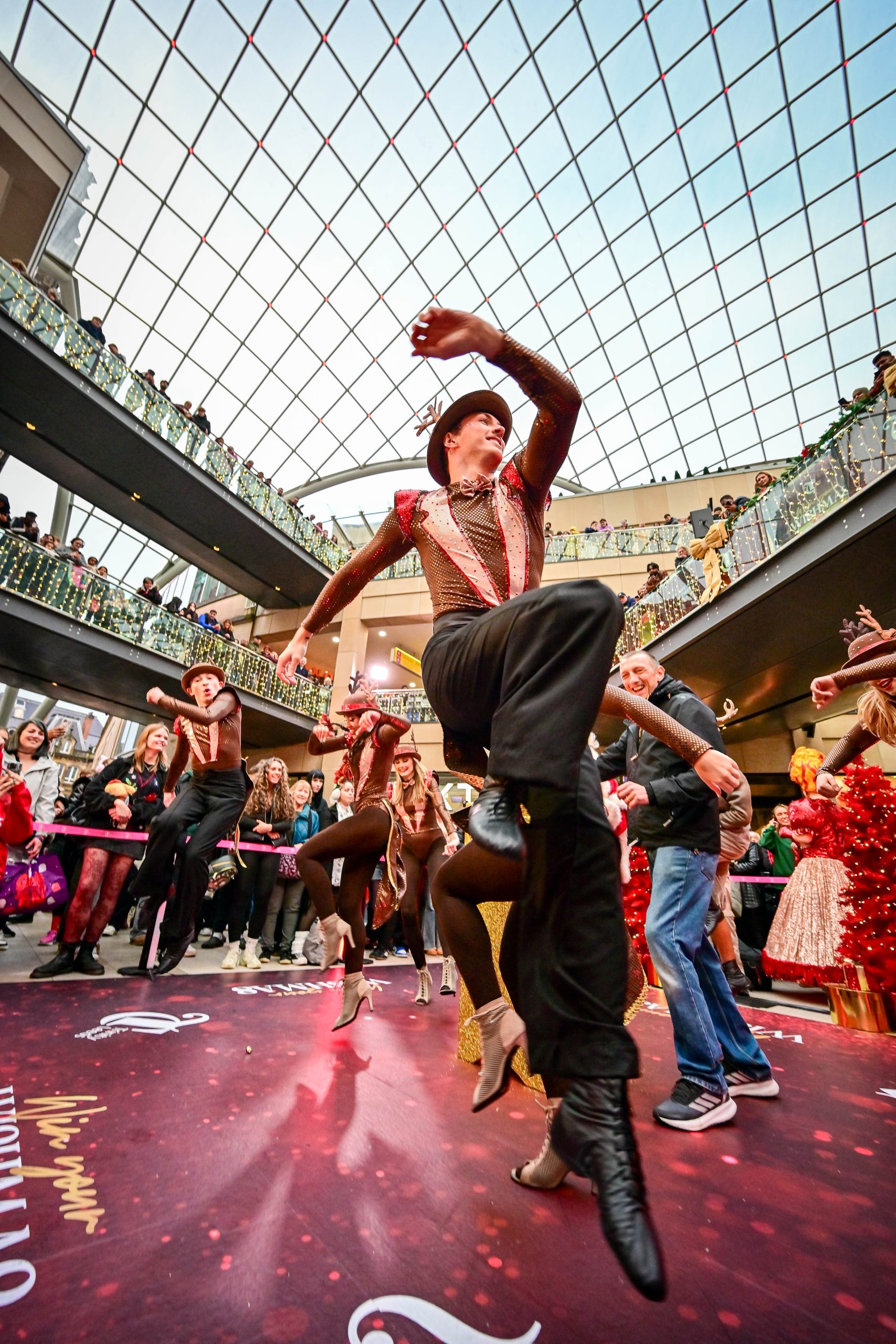 inside a glass roofed building where a few people in glittery costumes are dancing for a crowd